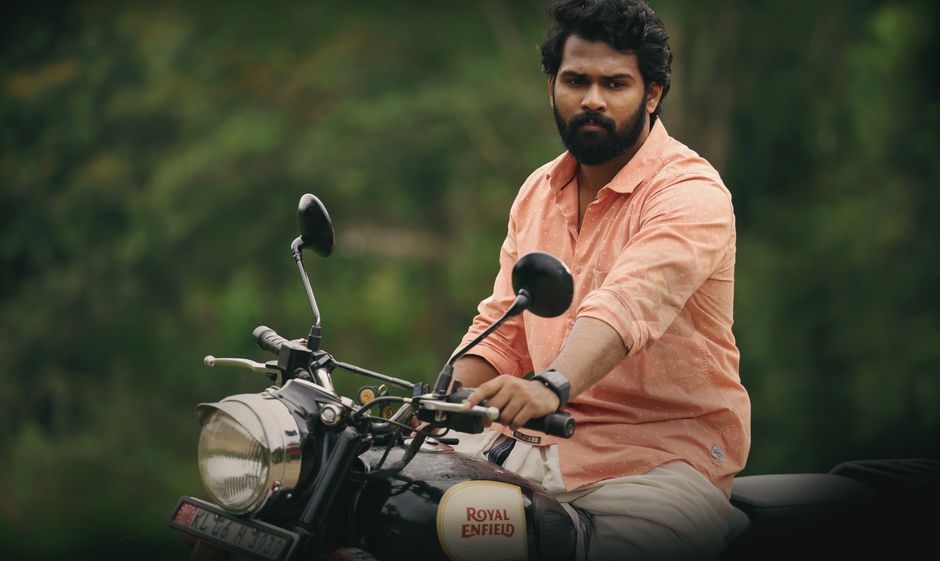 A man with dark hair and a beard, wearing an orange long-sleeve shirt and a wristwatch, is sitting on a black Royal Enfield motorcycle outdoors. The background features blurred green foliage, indicating a natural environment. The motorcycle's chrome headlamp, side mirrors, and handlebar are visible, with the man gripping the handlebars and looking towards the camera. The scene is well-lit with natural daylight, and the setting suggests a casual ride or transport of personal belongings, relevant to house removals or moving services. This image conveys the process of transportation, possibly associated with packing and moving activities, as offered by Man with Van Enfield Wash.