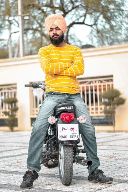 A man of South Asian descent wearing a cream turban and a yellow and white striped long-sleeve shirt is sitting on a black Bullet motorcycle parked on a paved area outdoors. His arms are crossed, and he has a beard. The motorcycle has a red tail light, a white license plate with the registration number PB46L6051, and a red camera mounted on the rear. In the background, there is a cream-colored building with balconies, metal railings, and two small, shaped bushes on either side of the motorcycle. The sky appears to be overcast, and several tall, leafless trees are visible behind the building. This image can support content related to transportation, packing, or moving logistics, relevant to house removal services provided by Man with Van Enfield Wash.