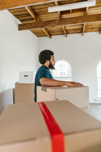A man with dark curly hair and a beard, wearing a teal T-shirt, is standing inside a room with a wooden ceiling and large arched windows allowing natural light. He is involved in a home relocation process, handling several cardboard boxes that are stacked and lined up on the floor and partially inside the room. The boxes vary in size and are sealed with packing tape; some are plain plain brown, while others are decorated with red tape. The room appears to be in the process of packing or unpacking, possibly during a house move, with the boxes ready for transportation. The setting suggests a professional removal service, such as Man with Van Enfield Wash, engaged in furniture transport and packing and moving activities as part of house removals near Enfield Town Station.