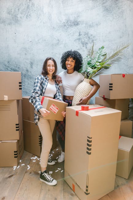 Two women inside a room surrounded by cardboard boxes, some sealed with red tape, indicating a home relocation process. The woman on the left, with shoulder-length brown hair, is wearing a checked shirt over a white top and light-colored trousers, holding a smaller box. The woman on the right, with curly black hair, is dressed in a white top and darker trousers, holding a large decorative vase with green palm leaves. The boxes are stacked and placed on a wooden floor, with some boxes positioned near an open doorway and others on the ground, suggesting the loading or unloading stage of furniture transport and packing during a house move. The background features a textured, light-colored wall, and the scene is illuminated with natural or soft lighting, reflecting a professional yet casual environment typical of moving and removal services offered by Man with Van Enfield Wash. This image visually represents packing, furniture transport, and the logistics involved in local house removals near Enfield Town Station.