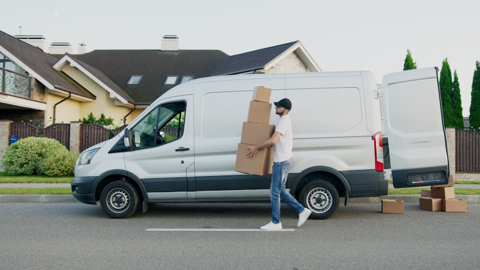 A man wearing a white t-shirt, blue jeans, white sneakers, and a black cap is loading three cardboard boxes into the side of a white cargo van parked on a residential street. One box is being carried towards the open rear door of the van, while two other boxes are placed on the ground nearby. The van is positioned parallel to the curb in front of modern houses with pitched roofs, some with skylights, and green trees and bushes are visible in the background. The scene is outdoors during daylight, with natural lighting highlighting the packing and loading process typical of home relocation or furniture transport services. Man with Van Enfield Wash specializes in removals, and this image depicts an active loading process involving packaging materials and the vehicle used for house moves.