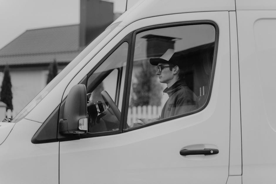 A man wearing glasses, a dark cap, and a jacket is sitting inside a white delivery van, visible through the closed passenger-side window. The van is parked outside a residential property with a driveway or street in front, as indicated by a backing fence and nearby houses. The interior of the van shows a vague glimpse of the dashboard and steering wheel. The background includes a house with a tiled roof and trees, suggesting a suburban setting. The man appears to be reviewing documents or a checklist, possibly related to a home relocation or moving logistics. The scene reflects a loading process or collection of packing materials, with the van ready for furniture transport or packing and moving tasks, and is associated with professional removals services offered by Man with Van Enfield Wash.